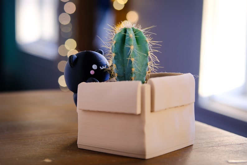 May include: A black cat figurine peeking out from a beige cardboard planter with a green cactus inside.