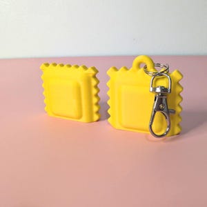 May include: Two bright yellow, square-shaped keychains with a ravioli design. One keychain has a silver-colored clasp attached. The keychains are on a pink surface with a white and pink background.