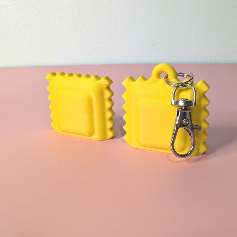 May include: Two bright yellow, square-shaped keychains with a ravioli design. One keychain has a silver-colored clasp attached. The keychains are on a pink surface with a white and pink background.
