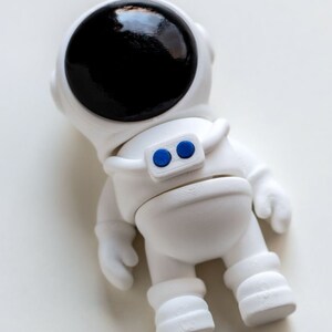 May include: White astronaut figurine with a black visor and blue buttons on the chest. The figurine is standing on a white surface.