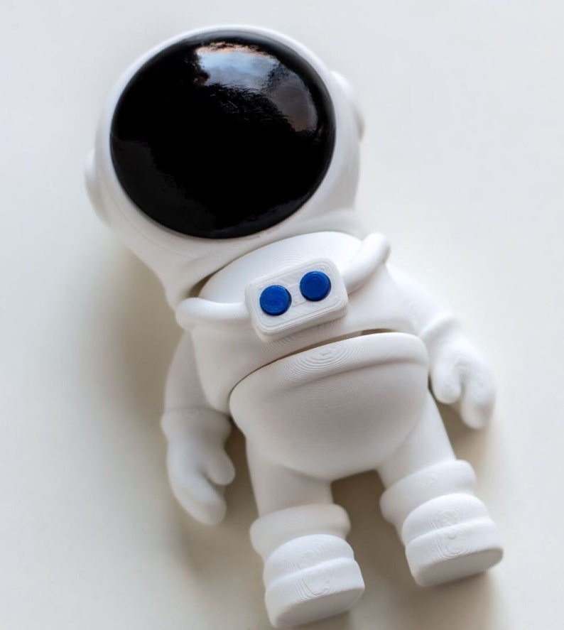 May include: White astronaut figurine with a black visor and blue buttons on the chest. The figurine is standing on a white surface.