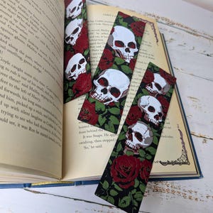 May include: Four decorative bookmarks with a skull and rose design. The bookmarks feature white skulls with red accents and red roses on a black background. The bookmarks are placed on an open book.