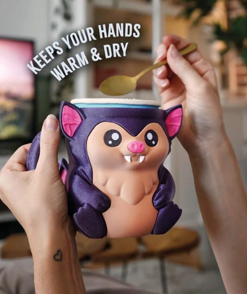 May include: A novelty mug shaped like a cartoon bat, with purple, pink, and tan colors. The mug has large eyes, a pink nose, and small fangs. Text on the image reads "KEEPS YOUR HANDS WARM & DRY". A gold spoon is held above the mug.