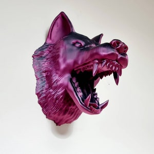 May include: A magenta and black wolf head sculpture with an open mouth, revealing sharp teeth. The sculpture is mounted on a white wall. The wolf's fur has a textured appearance.