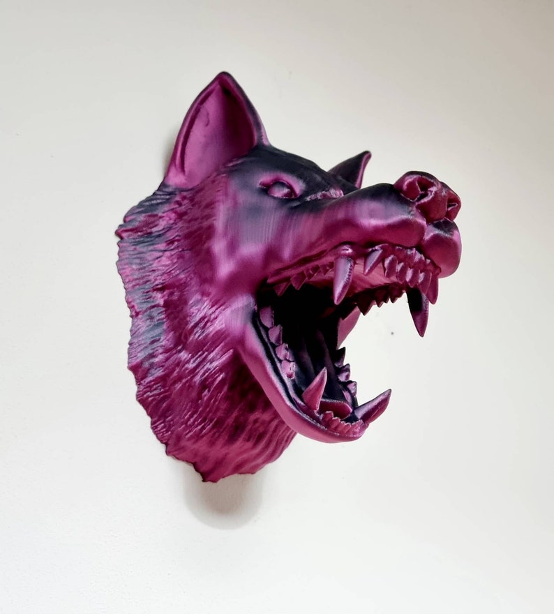 May include: A magenta and black wolf head sculpture with an open mouth, revealing sharp teeth. The sculpture is mounted on a white wall. The wolf's fur has a textured appearance.