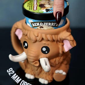 May include: A brown mammoth-shaped ice cream pint holder with a 92 mm opening. The holder has large black eyes, pink ears, and white tusks. A pint of Ben & Jerry's ice cream is partially visible inside the holder.