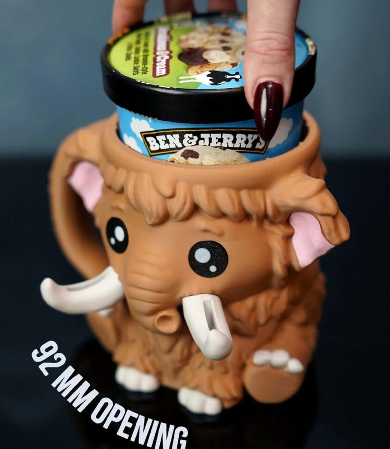May include: A brown mammoth-shaped ice cream pint holder with a 92 mm opening. The holder has large black eyes, pink ears, and white tusks. A pint of Ben & Jerry's ice cream is partially visible inside the holder.