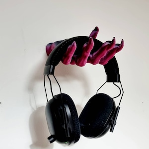 Puede incluir: Un auricular negro con un micr&oacute;fono colgando de un soporte para auriculares en forma de mano impreso en 3D. La mano est&aacute; pintada en tonos de rosa y morado.