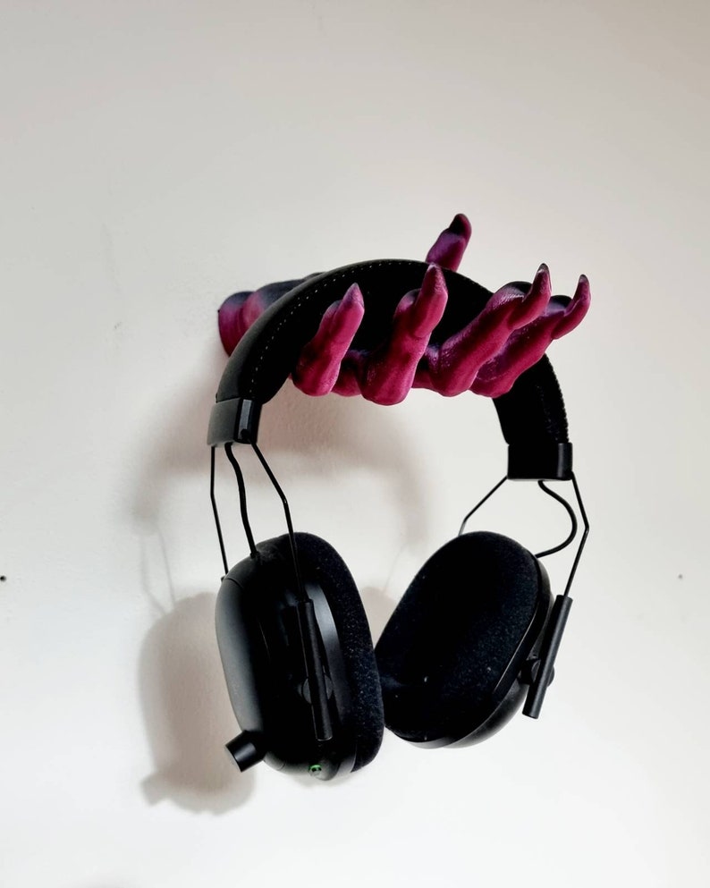 Puede incluir: Un auricular negro con un micr&oacute;fono colgando de un soporte para auriculares en forma de mano impreso en 3D. La mano est&aacute; pintada en tonos de rosa y morado.