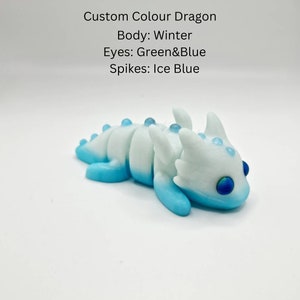 May include: A small, white and blue dragon figurine with green and blue eyes. The dragon has a winter white body with blue accents and ice blue spikes.