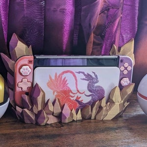 May include: A 3D printed Nintendo Switch stand featuring a purple and gold crystal design. The stand has a cutout for the Switch console and a space for the Joy-Cons. The stand is decorated with a purple and red dragon design.