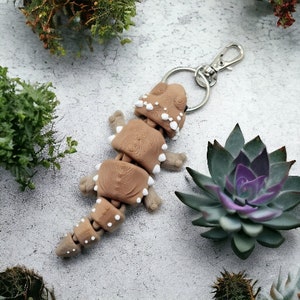 May include: A brown and white polymer clay keychain shaped like a lizard with a silver key ring. The lizard has a segmented body and white spots.