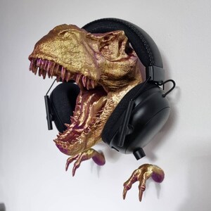 May include: A gold and purple 3D printed dinosaur head with a pair of black headphones hanging from it. The dinosaur's mouth is open and its teeth are visible. The dinosaur's claws are also visible.