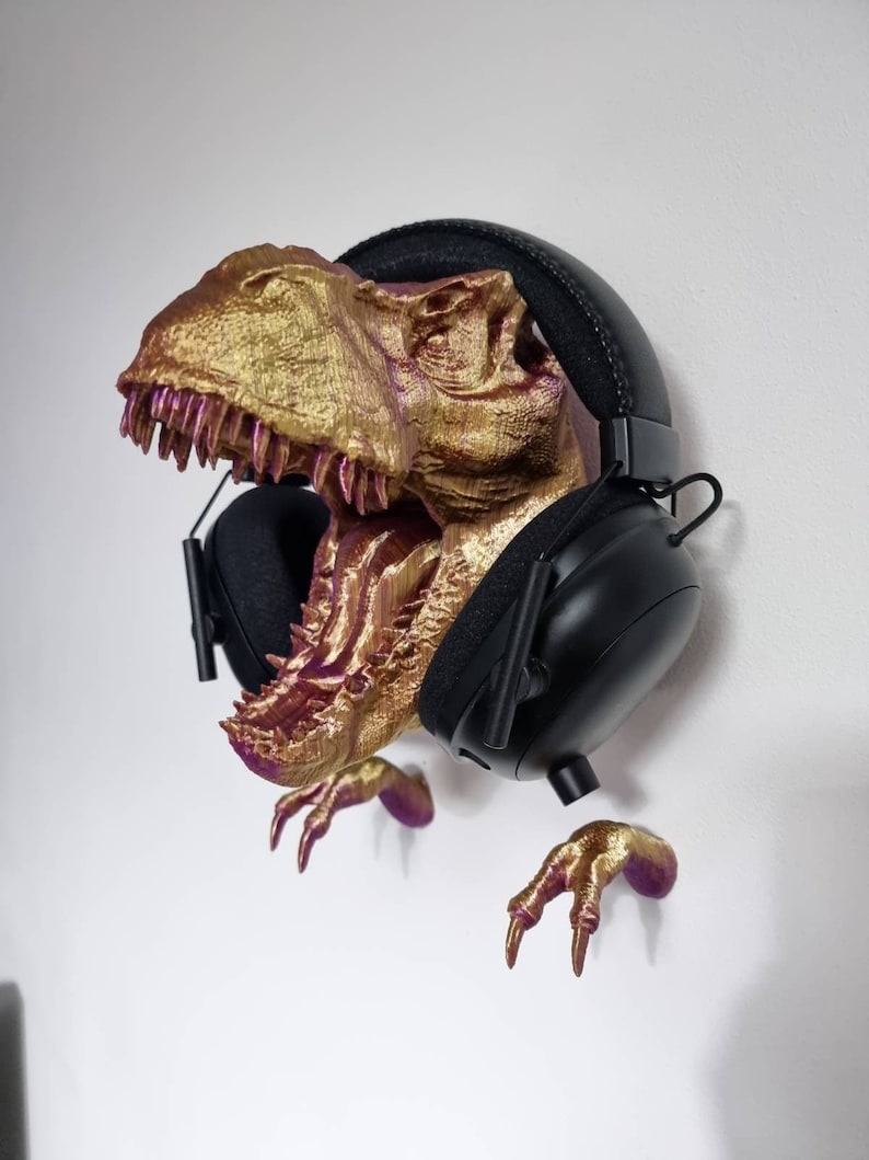 May include: A gold and purple 3D printed dinosaur head with a pair of black headphones hanging from it. The dinosaur's mouth is open and its teeth are visible. The dinosaur's claws are also visible.