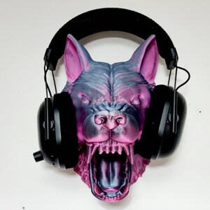 May include: A 3D-printed wolf head in shades of gray and pink, with an open mouth and visible teeth. Black headphones are placed over the wolf's head, resting on the ears. The wolf head is mounted on a white wall.