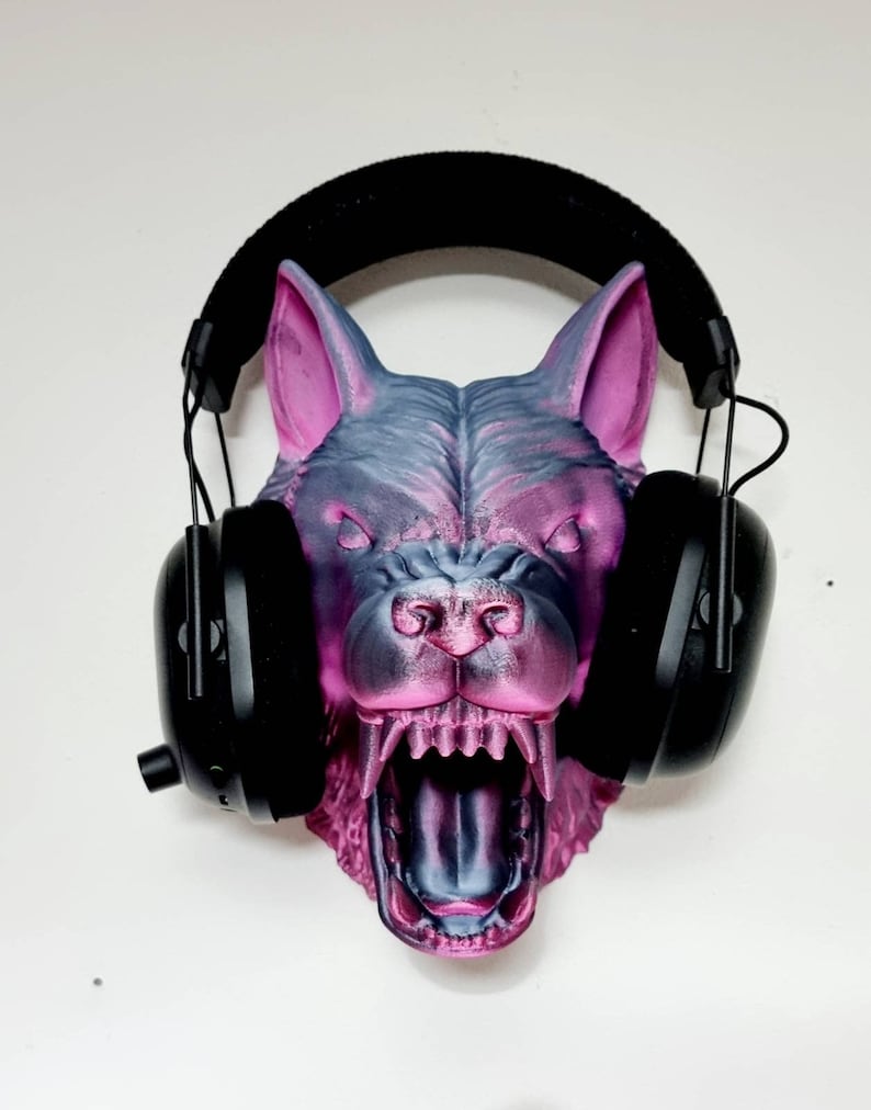May include: A 3D-printed wolf head in shades of gray and pink, with an open mouth and visible teeth. Black headphones are placed over the wolf's head, resting on the ears. The wolf head is mounted on a white wall.