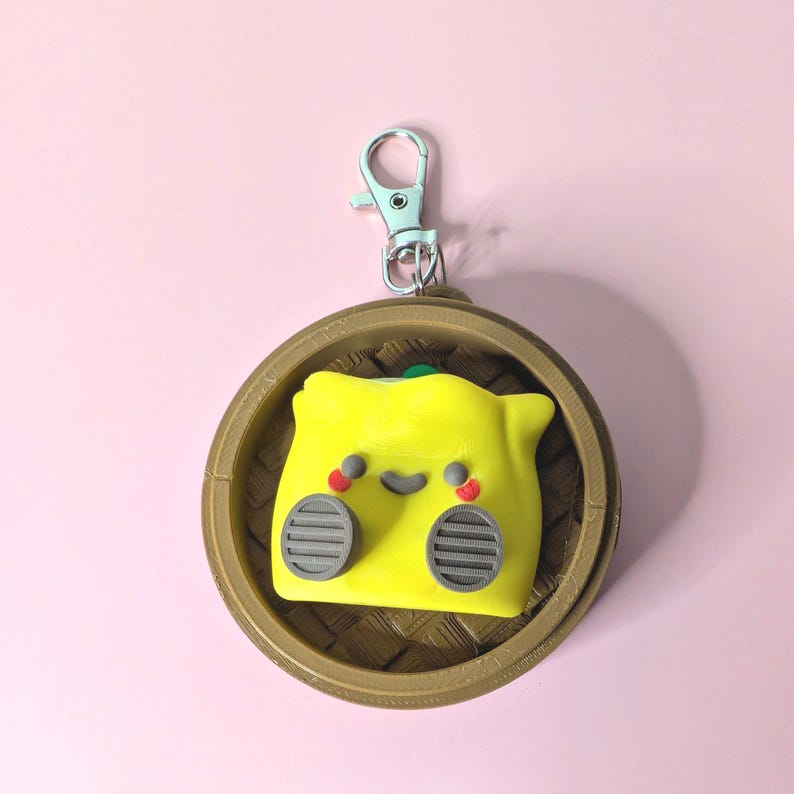May include: A yellow dumpling-shaped keychain with a smiling face, red cheeks, and gray details, set in a brown bamboo steamer. The keychain has a silver clasp and a green accent on top. The background is a solid pink.