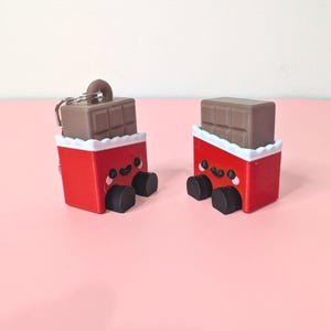 Puede incluir: Dos llaveros de barra de chocolate novedosos. Cada llavero presenta un envoltorio rojo con una cara sonriente, pies negros y una barra de chocolate marr&oacute;n en la parte superior. Los envoltorios tienen un borde blanco festoneado.