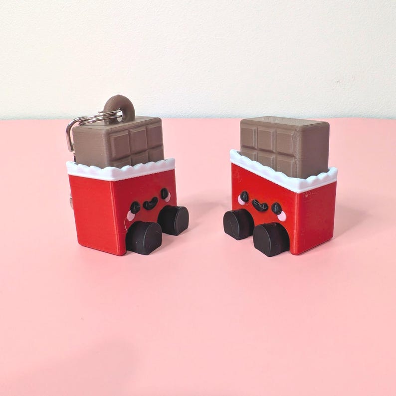 Puede incluir: Dos llaveros de barra de chocolate novedosos. Cada llavero presenta un envoltorio rojo con una cara sonriente, pies negros y una barra de chocolate marr&oacute;n en la parte superior. Los envoltorios tienen un borde blanco festoneado.