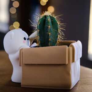 May include: A white ceramic figurine with a cute face is holding a brown cardboard planter with a green cactus inside.