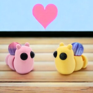 May include: Two small, colorful, 3D printed figurines shaped like caterpillars. The pink caterpillar has a purple and pink shell and black eyes. The yellow caterpillar has a purple shell and black eyes. The figurines are on a wooden surface.
