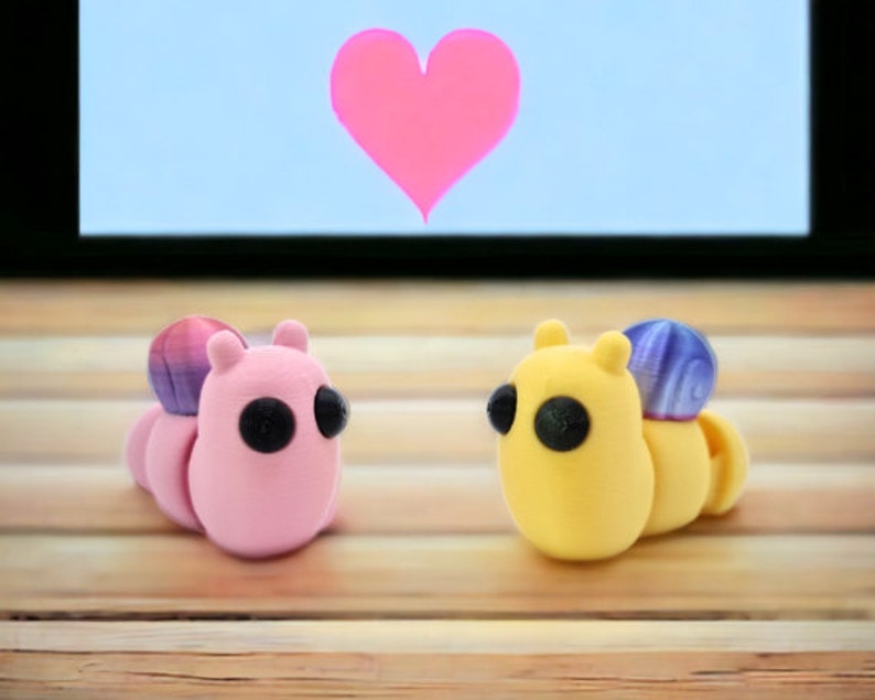 May include: Two small, colorful, 3D printed figurines shaped like caterpillars. The pink caterpillar has a purple and pink shell and black eyes. The yellow caterpillar has a purple shell and black eyes. The figurines are on a wooden surface.