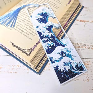 May include: A rectangular bookmark featuring a stylized depiction of a large ocean wave in shades of blue and white. The bookmark has a blue tassel attached. The bookmark is placed in an open book.