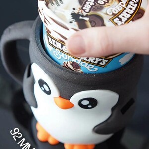 May include: A black, white, and orange penguin-shaped ice cream pint holder. The holder has a 92 mm wide opening and is holding a pint of Ben & Jerry's ice cream. The ice cream container has the words "Cookie Vermont-ster" on the lid.