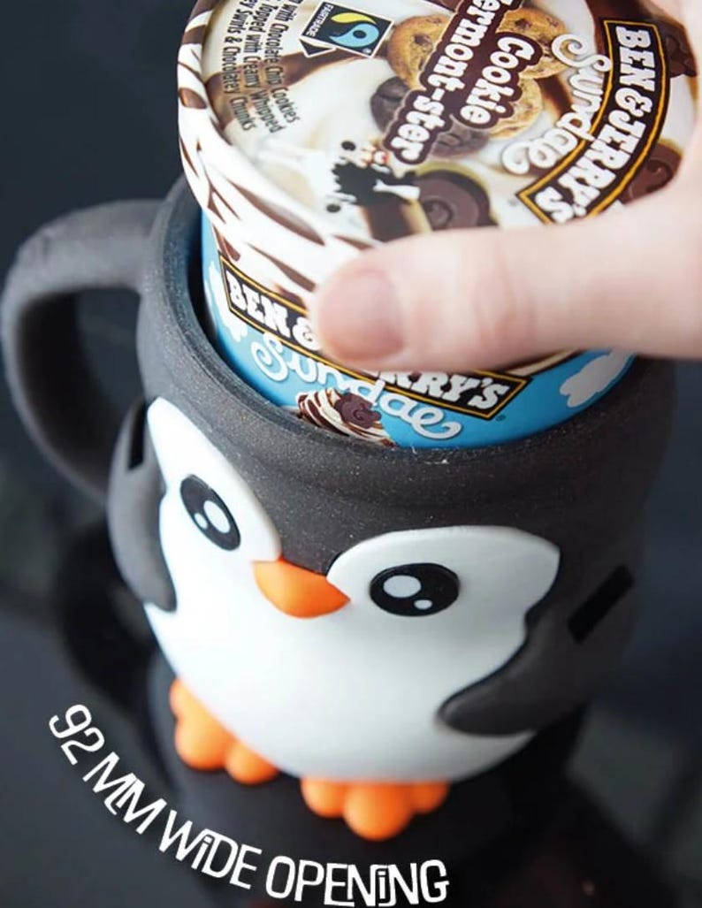 May include: A black, white, and orange penguin-shaped ice cream pint holder. The holder has a 92 mm wide opening and is holding a pint of Ben & Jerry's ice cream. The ice cream container has the words "Cookie Vermont-ster" on the lid.
