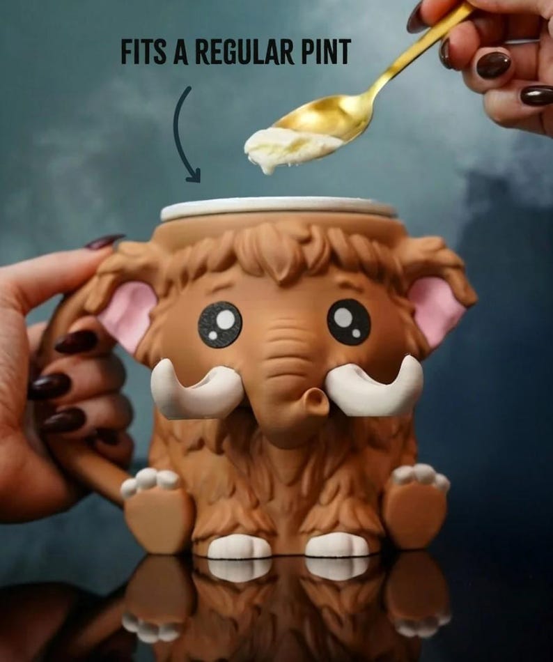 May include: A brown mammoth-shaped mug with large tusks, ears, and feet. The mug has a handle and is being filled with ice cream from a gold spoon. Text above the mug reads "FITS A REGULAR PINT".