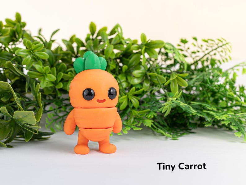 May include: A small, orange carrot-shaped figurine with a green leafy top and a smiling face. The figurine is made of clay and has a cartoonish appearance. The figurine is sitting on a white surface with green foliage in the background. The text "Tiny Carrot" is written below the figurine.