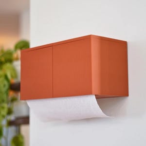 May include: A wall-mounted paper towel dispenser in a warm orange hue. The dispenser has a rectangular shape with a textured, ribbed surface. White paper towels are partially dispensed from the bottom. The dispenser is mounted on a white wall.