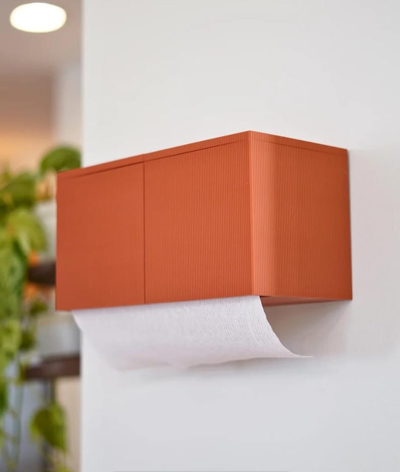 May include: A wall-mounted paper towel dispenser in a warm orange hue. The dispenser has a rectangular shape with a textured, ribbed surface. White paper towels are partially dispensed from the bottom. The dispenser is mounted on a white wall.