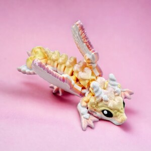 May include: A small, hand-painted dragon figurine with a white, pink and yellow colour scheme. The dragon is posed in a playful, curled position with its wings spread out.