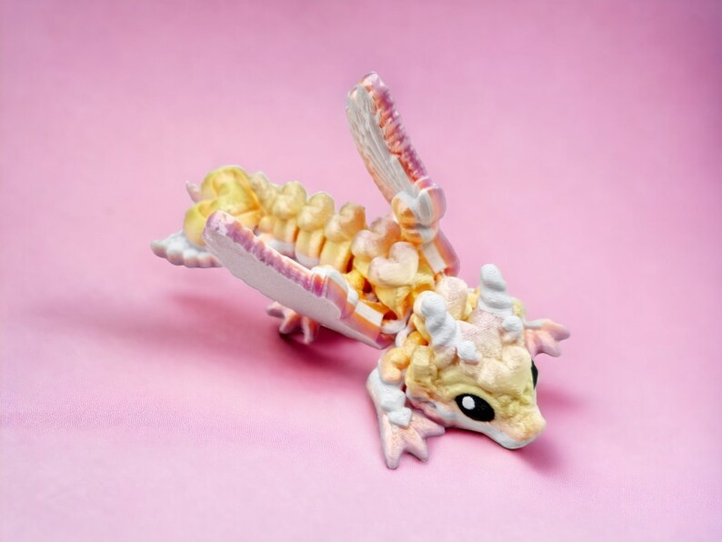 May include: A small, hand-painted dragon figurine with a white, pink and yellow colour scheme. The dragon is posed in a playful, curled position with its wings spread out.