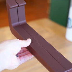 Peut inclure: Objet brun en forme de L avec une section transversale rectangulaire. L'objet est tenu dans la main d'une personne. Il a un dessus plat et une rainure au centre du c&ocirc;t&eacute; long. L'objet semble &ecirc;tre en bois ou dans un mat&eacute;riau similaire.