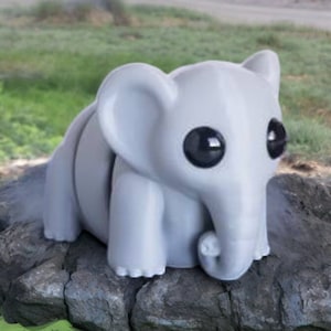 May include: A small, gray, 3D printed elephant figurine with large black eyes. The elephant is sitting on a gray rock.