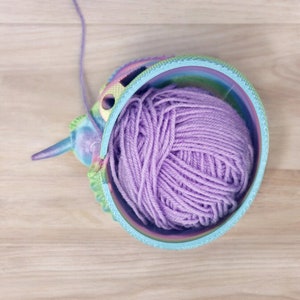 May include: A colorful yarn bowl with a purple ball of yarn inside. The bowl is shaped like a unicorn head and has a rainbow design.