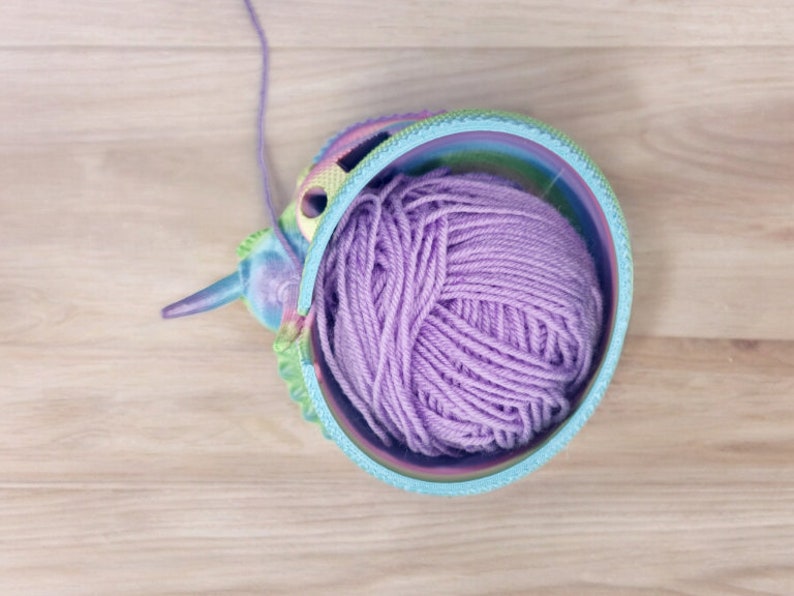 May include: A colorful yarn bowl with a purple ball of yarn inside. The bowl is shaped like a unicorn head and has a rainbow design.