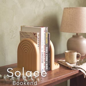 Op de afbeelding: Beige boogvormige boekensteunen met een gelaagd ontwerp, die boeken op een houten oppervlak vasthouden. Het woord "Solace" staat op de voorkant. Een lamp en een mok staan op de achtergrond.