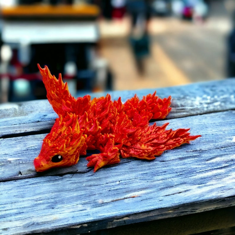 May include: A small, red and orange figurine of a mythical creature with a long tail and wings. The creature is made of a textured material that resembles flames.