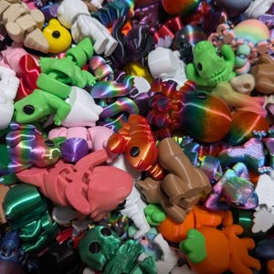 Peut inclure: Un ensemble de jouets imprim&eacute;s en 3D aux couleurs vives, dont des grenouilles, des astronautes et des formes abstraites. Les jouets sont de diff&eacute;rentes couleurs, notamment vert, orange, rose et arc-en-ciel. Gros plan des jouets.