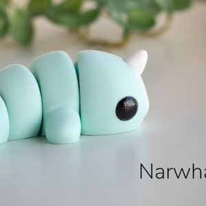May include: A light blue, segmented, narwhal figurine with black eyes and a white horn. The figurine is lying on a white surface. "Narwhal" is written in black text below the figurine.
