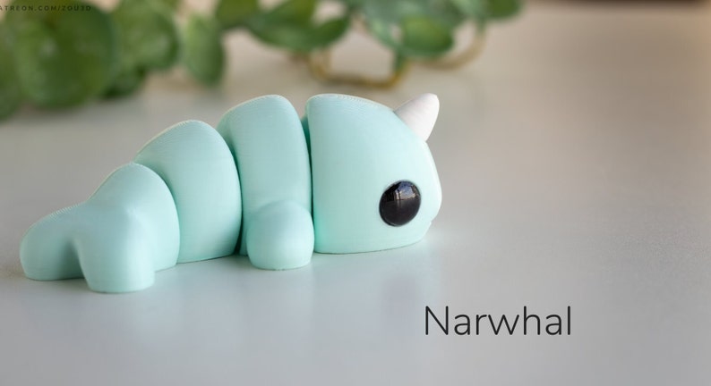 May include: A light blue, segmented, narwhal figurine with black eyes and a white horn. The figurine is lying on a white surface. "Narwhal" is written in black text below the figurine.