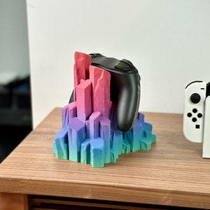 May include: A 3D printed crystal-shaped stand in shades of pink, purple, and blue holds a black video game controller. The stand is on a wooden surface.