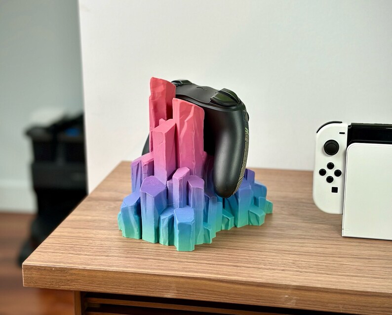 May include: A 3D printed crystal-shaped stand in shades of pink, purple, and blue holds a black video game controller. The stand is on a wooden surface.