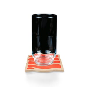 May include: A tall, clear glass with a black interior sits on a wooden coaster with a wavy orange design. The coaster is rectangular with rounded edges. The glass is empty and has a simple, modern design.