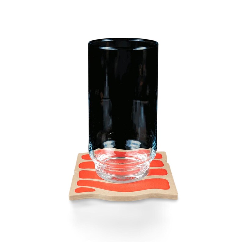 May include: A tall, clear glass with a black interior sits on a wooden coaster with a wavy orange design. The coaster is rectangular with rounded edges. The glass is empty and has a simple, modern design.