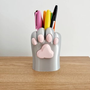 May include: A silver paw-shaped desk organizer with pink paw prints holds a variety of pens and markers.