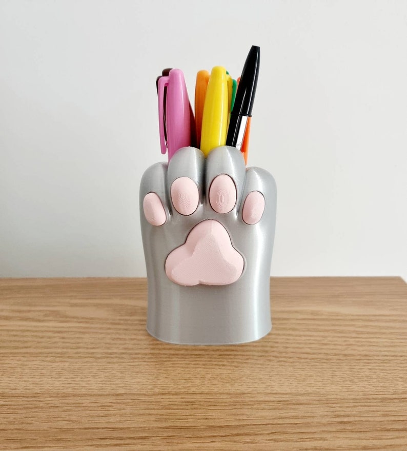 May include: A silver paw-shaped desk organizer with pink paw prints holds a variety of pens and markers.